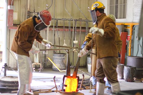 people in foundry gear hold large glowing crucible with metal tongs in foundry with equipment and gear all around 